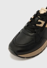 Svart sneaker laget av glatt skinn med gullakkenter. Har lisse, rund tå og en beige gummisåle. Teksturert søm gir detaljer.