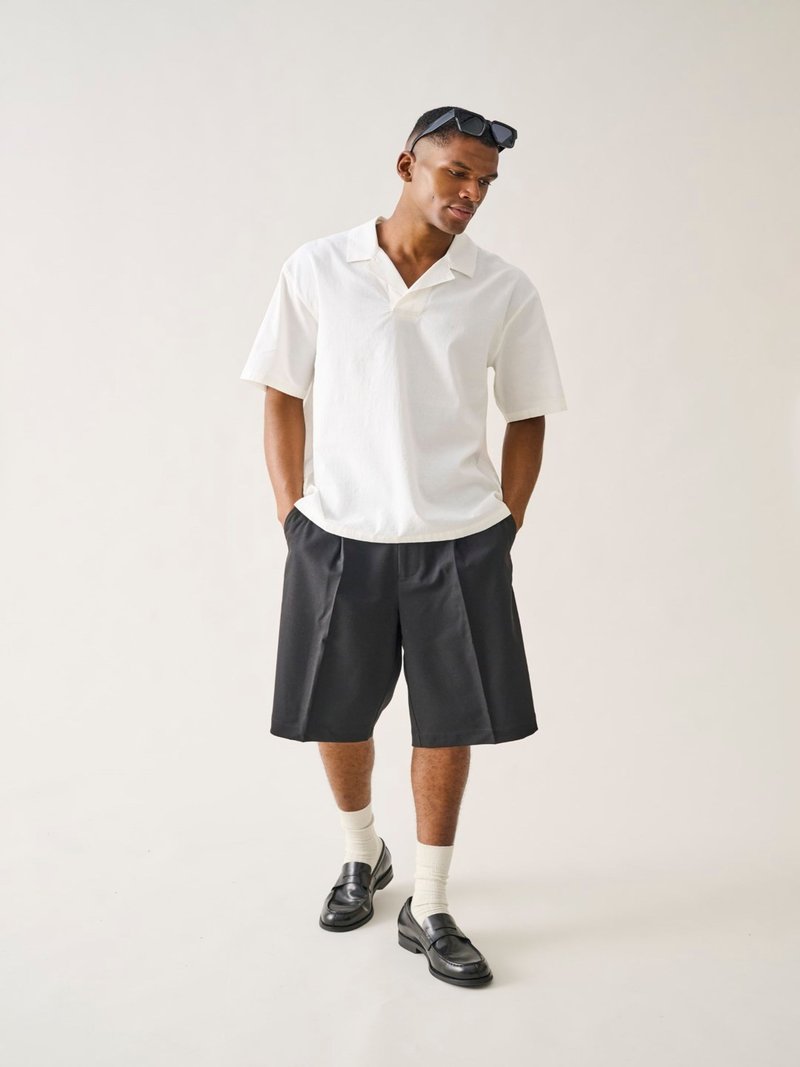 Homme portant une chemise blanche à manches courtes avec col, un short noir jusqu'aux genoux, des chaussettes blanches, des mocassins noirs et des lunettes de soleil sur la tête, debout les mains dans les poches.
