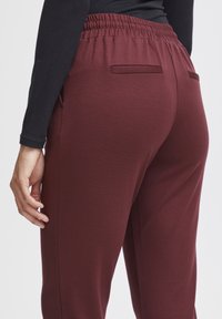Burgundy joggers featuring an elastic waistband, two back pockets, and a smooth texture, shown from the back with a black long-sleeve top.