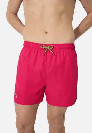 Hombre vistiendo pantalones de baño rosa brillante con un cordón verde, de pie con las manos detrás de la espalda contra un fondo liso.