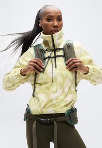 Ανοιχτό κίτρινο φλις μπλουζάκι με καμουφλάζ μοτίβο, ψηλό γιακά και μαύρο φερμουάρ· πράσινοι ιμάντες ιμάντα ασφαλείας και ζώνη χρηστικότητας με κλιπ και τσέπες.