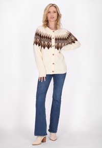 Cardigan crema con motivo geometrico marrone, bottoni in legno e maniche lunghe, abbinato a jeans a zampa d'elefante blu e stivali con tacco beige.
