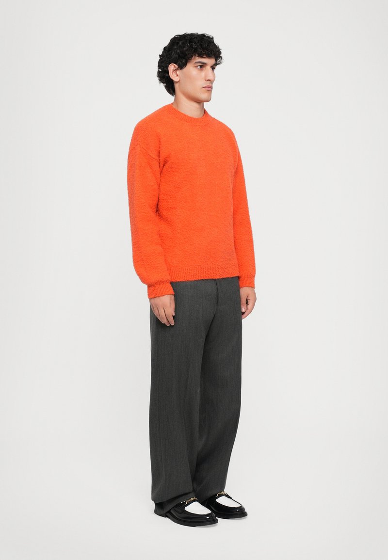 Uomo che indossa un luminoso maglione arancione fluffy, pantaloni grigi scuri larghi e mocassini in bianco e nero, in piedi contro uno sfondo bianco semplice.