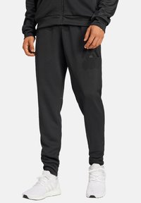 Zwarte joggingbroek met een zachte textuur en een taps toelopende pasvorm, met een subtiele logo-accent. Gecombineerd met witte sportschoenen.