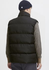 Schwarze Steppweste mit hohem Kragen, mit vertikalen Steppnähte. Trägt sie über einem strukturierten grauen Pullover und einer gerippten marineblauen Mütze.