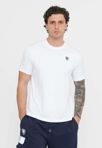 Maglietta in cotone bianco con collo rotondo e maniche corte, caratterizzata da un piccolo logo nero sul petto. Tessuto liscio e design semplice.