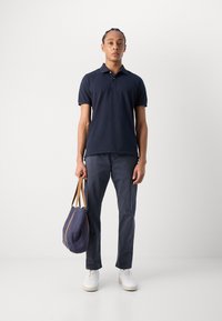 Polo shirt marine à manches courtes, associé à un pantalon foncé et à des baskets blanches. Le modèle tient un sac marine avec des poignées brunes.