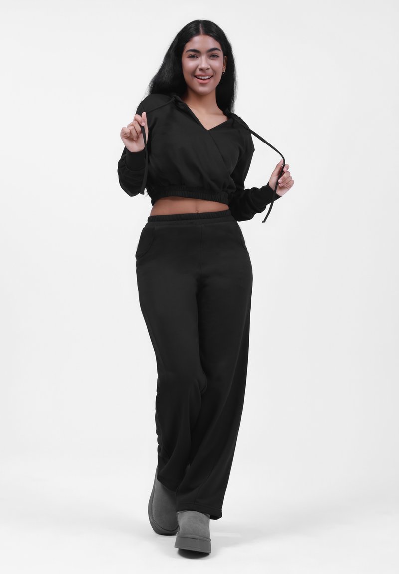 Sudadera negra corta con cordón, combinada con pantalones negros holgados. El modelo lleva calzado gris, presentado sobre un fondo blanco.