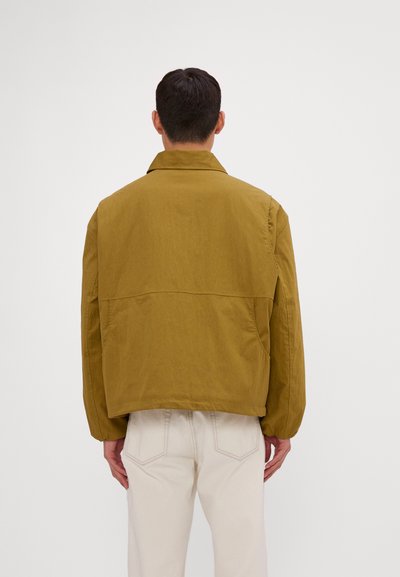 Jacheta oversized de culoare khaki cu guler plat și manșete elastice. Prezintă o textură netedă și o croială pătrată. Vederea din spate nu arată închideri vizibile.