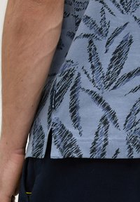 Chemise à manches courtes en tissu bleu clair avec un motif de feuilles en noir et bleu foncé. Détails de fente sur le côté et étiquette de logo visibles.