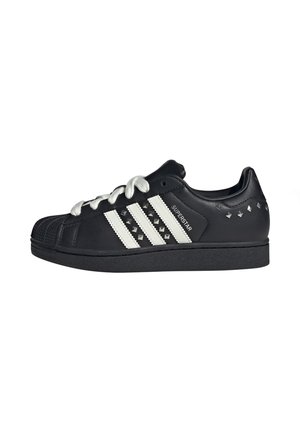 Zapatilla Adidas Superstar negra de corte bajo con cordones blancos, tres franjas blancas con tachuelas negras y tachuelas metálicas en el cuello del talón.