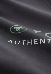 Donkergrijze t-shirt met groen en witte opdruk. De tekst bevat een logo en het woord "AUTHENTIC." Gladde stoftextuur.