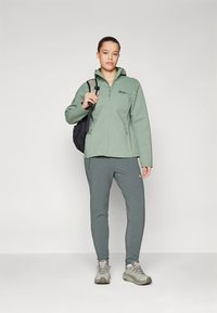 Gaiši zaļa softshell jaka ar rāvējslēdzēja aizdari, kopā ar atbilstošām sporta biksēm un pelēkām pārgājienu kurpēm. Mugursoma nēsāta uz kreisā pleca.