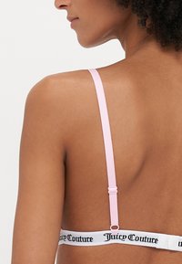 Rozā regulējamā siksniņa krūšturis ar baltu joslu, uz kuras ir melns "Juicy Couture" teksts, uzsverot elastīgu materiālu un minimālistisku dizainu.