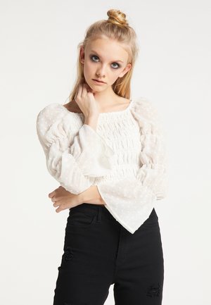 Weiße Bluse mit Puffärmeln, mit geraffter Textur und Punkte-Muster, kombiniert mit schwarzen, verwaschenen Jeans. Die Bluse hat einen weiten Ausschnitt.