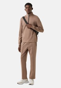 Bruine sweatvest met rits en een gladde textuur, gecombineerd met bijpassende taps toelopende broek. Beide hebben een minimalistisch ontwerp en subtiele stiksel details.