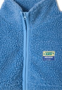Chaqueta de polar azul claro con cierre de cremallera, que presenta una superficie texturizada y suave, y una etiqueta con detalles en verde y amarillo.