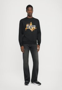 Μαύρος φούτερ με το λογότυπο "VERSACE JEANS COUTURE" σε λευκά και πορτοκαλί φλόγες; συνδυασμένο με σκούρες γκρι τζιν και μαύρα αθλητικά παπούτσια.