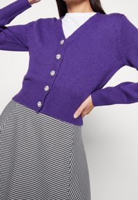 Lila gestrickte Strickjacke mit V-Ausschnitt, geripptem Saum und fünf blumenförmigen Knöpfen. Kombiniert mit einem schwarz-weißen Hahnentrittmuster-Rock.