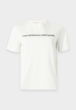 Femme debout, une main dans la poche, portant un T-shirt blanc avec un texte et un pantalon beige à larges jambes avec des talons hauts marron.