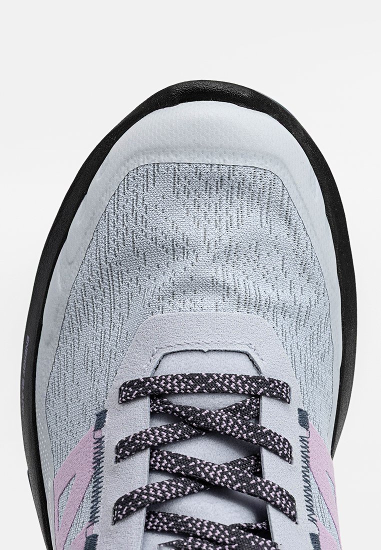 Acercamiento de la punta de una zapatilla gris y rosa con cordones negros estampados y parte superior de tela texturizada, enmarcada por un borde de suela negra.