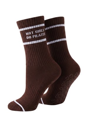 Nogi w brązowych legginsach w ciemnobrązowych skarpetkach ze białymi paskami i napisem, jedna stopa płasko, druga wysunięta na jasnym tle.