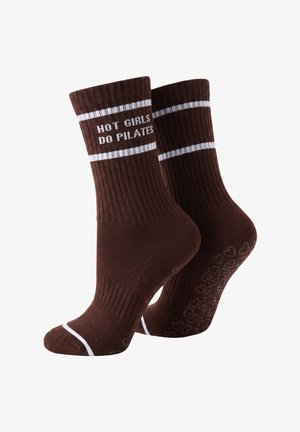 Nogi w brązowych legginsach w ciemnobrązowych skarpetkach ze białymi paskami i napisem, jedna stopa płasko, druga wysunięta na jasnym tle.