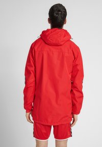 Rød vandtæt jakke med hætte, der har lange ærmer og en lige pasform. Ingen synlige lukninger eller mønstre. Bæres med røde shorts.