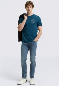 Modra bombažna majica z okrolim ovratnikom in okroglim tiskanim logotipom na prsih, kombinirana z modrimi kavbojkami in belimi supergami, zraven pa črna jakna prevrnjena čez ramo.