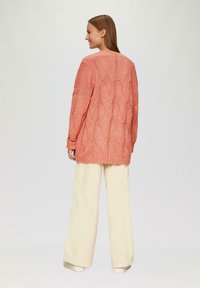 Helle korallenfarbene Strickjacke mit strukturiertem Muster, lockere Passform und langen Ärmeln, kombiniert mit weiten weißen Hosen.