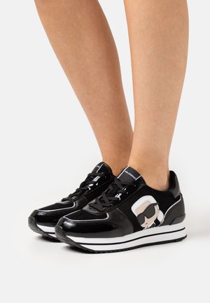Baskets noires et blanches avec un visage de dessin animé portant des lunettes de soleil sur les côtés, portées pieds nus sur un fond uni.