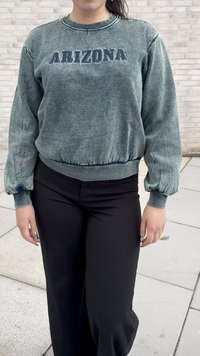 Persoană îmbrăcată cu un pulover verde tern "ARIZONA" și pantaloni negri, stând pe un paviment cu dale de culoare deschisă.