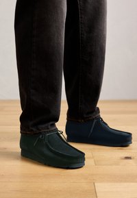 Person wearing black jeans and dark green suede moccasin shoes with laces, standing on a light wooden floor.