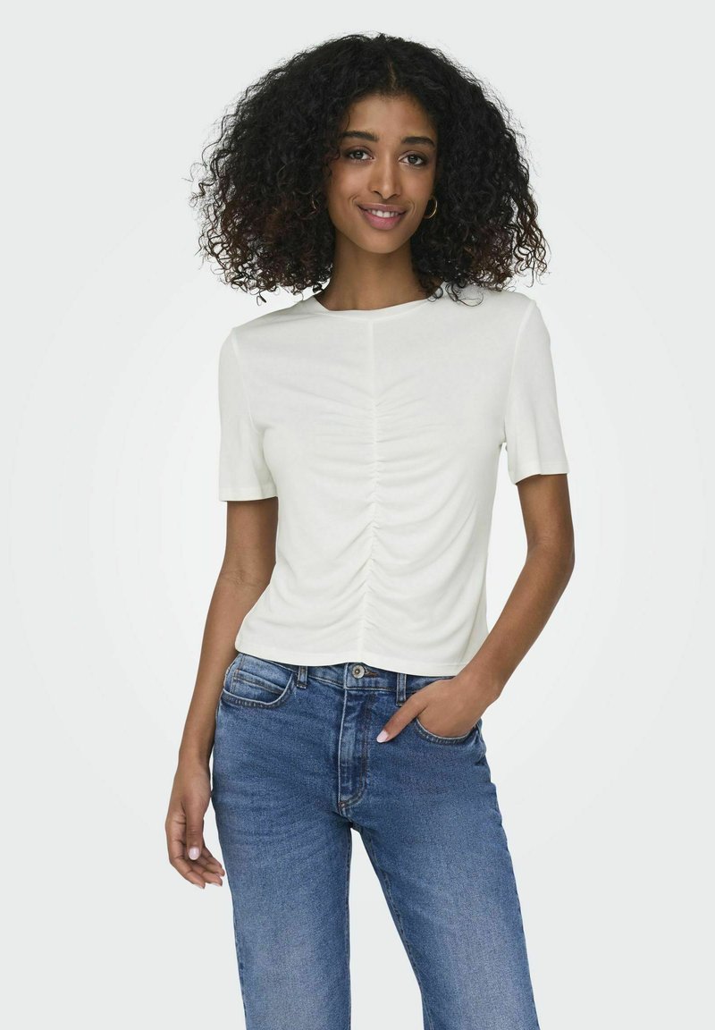 Top corto blanco de manga corta con detalles fruncidos en el centro y un escote redondo, combinado con jeans de mezclilla azul. Textura de tela suave.