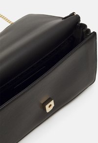 Czarna skórzana torebka o wyraźnie zarysowanym kształcie, ze złotym kwadratowym okuciem oraz wewnętrzną kieszenią na zamek. W tle widoczny jest łańcuszkowy pasek.