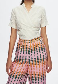 Blouse blanche texturée à nouer avec manches courtes ; associée à un pantalon rose orné d'un motif de pois verts et orange. Coupe décontractée, taille élastique.