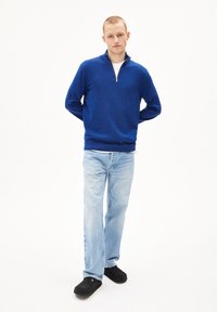 Pull bleu en tricot avec col zippé, associé à un jean bleu clair et des mocassins noirs, sur fond blanc.