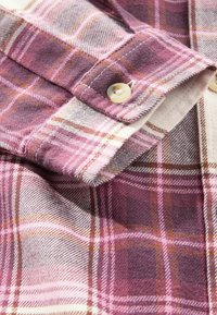 Détail d'une chemise en flanelle à carreaux violets et roses avec des accents blancs et beiges, présentant une patte de bouton et une texture douce.