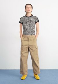 Síťový top s leopardím potiskem a černým lemem, spárovaný s béžovými cargo kalhotami s bočními kapsami. Model má na sobě jasně žluté tenisky.