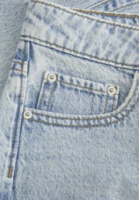 Ljusblå denimtyg har en liten, rektangulär ficka med silverdetaljer, synlig sömnad i en kontrasterande gul färg och en strukturerad yta.