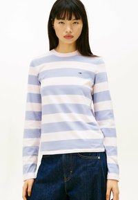 Camisa de rayas de manga larga en algodón de color azul claro y rosa, con un cuello redondo y un pequeño logo en el pecho. Combinada con jeans oscuros.