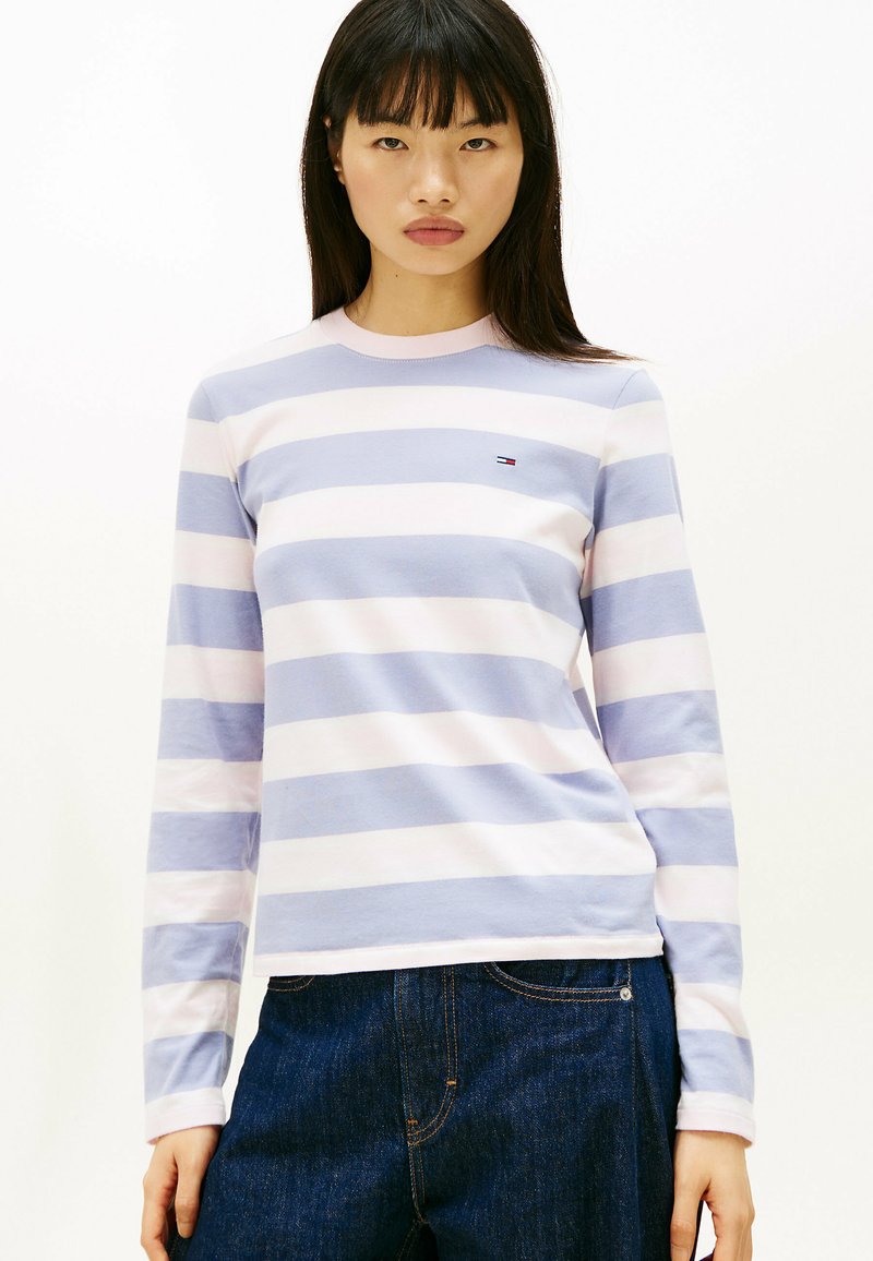 Camisa de rayas de manga larga en algodón de color azul claro y rosa, con un cuello redondo y un pequeño logo en el pecho. Combinada con jeans oscuros.