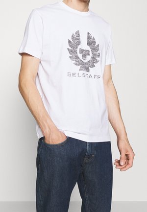 Mann trägt weißes T-Shirt mit kurzen Ärmeln und verblasstem schwarzem Belstaff-Logo sowie dunkelblaue Jeans, Hand in der Tasche, vor einem schlichten Hintergrund.