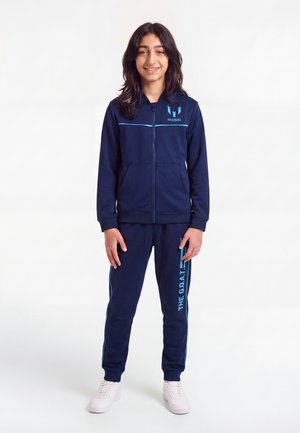 Enfant aux longs cheveux foncés se tient debout en souriant, portant un survêtement Messi bleu marine avec des baskets blanches sur un fond blanc uni.