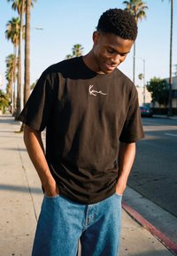 Joven con camiseta negra con diseño gráfico y vaqueros azules parado en una acera soleada con palmeras, con las manos en los bolsillos, mirando hacia abajo y sonriendo.
