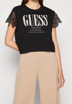 Sort bomulds-T-shirt med korte ærmer i blonder, med stort "GUESS" logo og tekst. Cropped design med løs pasform.