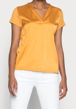Vrouw draagt een mosterdgele V-hals blouse met korte mouwen en witte broek, staand tegen een effen lichte achtergrond.