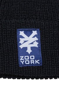 Čierna pletená čiapka s obdĺžnikovým modrým pásom, na ktorom je biele logo a text "ZOO YORK" na spodnej strane. Viditeľný detail textúrovaného materiálu.