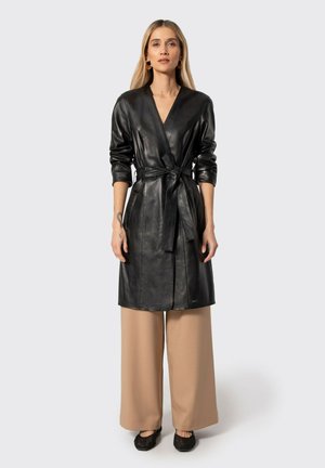 Donna che indossa un lungo cappotto in pelle nero con cintura sopra pantaloni beige a gamba larga e scarpe nere basse, in piedi davanti a uno sfondo chiaro e semplice.