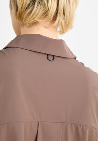 Top marrone con colletto e dettaglio plissettato sul retro, caratterizzato da una chiusura a loop nera al collo. Tessuto liscio e leggero.